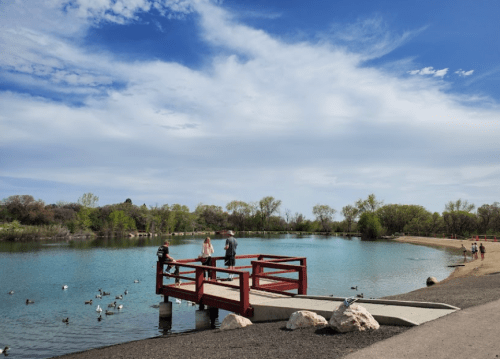 A serene lake scene with people on a red dock, surrounded by trees and ducks under a blue sky with clouds.
