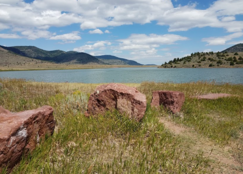 A serene landscape featuring a lake surrounded by mountains, with large red rocks in the foreground and grassy areas.