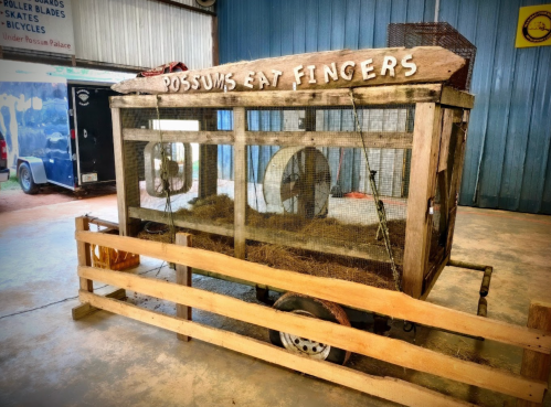 A wooden display trailer with a sign reading "Possums Eat Fingers," featuring a fenced area and a wheel inside.