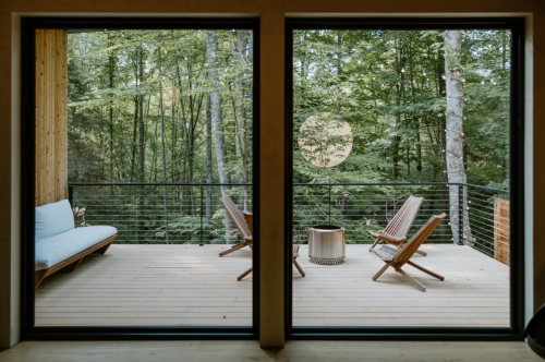 A serene outdoor deck with two wooden chairs and a fire pit, surrounded by lush green trees.