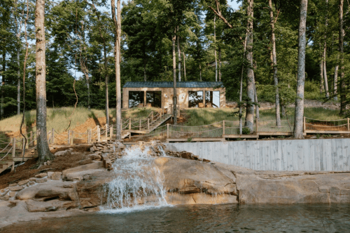 A rustic cabin by a small waterfall, surrounded by trees and grassy terrain.