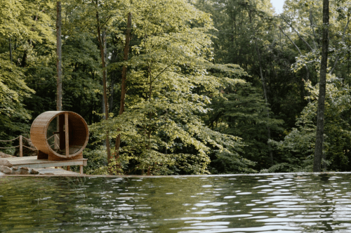 A serene forest scene featuring a wooden barrel sauna by a calm water body, surrounded by lush green trees.