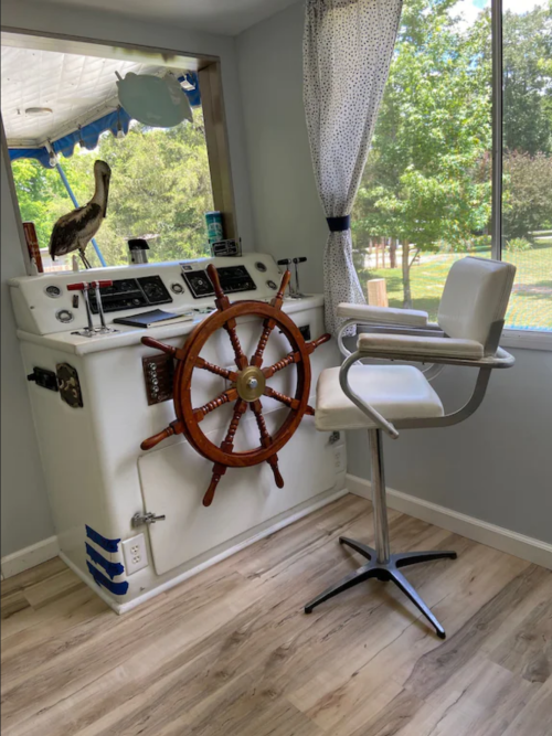 A unique room featuring a ship's wheel, a bar stool, and a window with a view of nature outside.