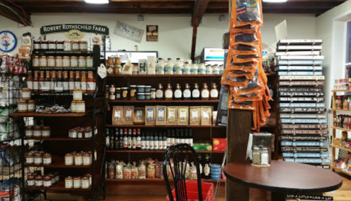 A cozy store interior featuring shelves of various jars, sauces, and snacks, with a small table in the foreground.