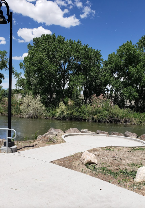 A serene riverside scene with trees, a pathway, and a clear blue sky dotted with clouds.