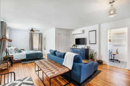 A bright, modern living space featuring a blue sofa, wooden floors, a bed, and a small desk area.