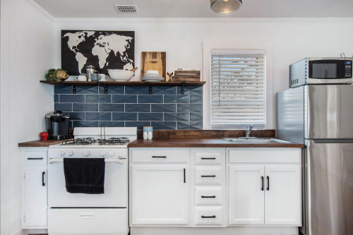 A modern kitchen with a gas stove, stainless steel fridge, blue tile backsplash, and open shelving with dishes.
