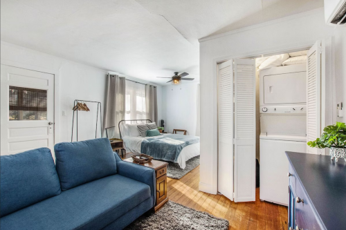 Cozy studio apartment with a blue sofa, bed, and laundry area, featuring natural light and simple decor.
