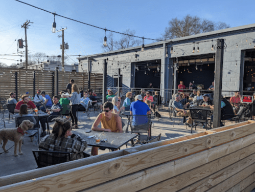 A lively outdoor patio filled with people enjoying drinks and food on a sunny day, with string lights overhead.