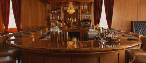 A warm, wooden bar with a chandelier, bottles, and glasses, featuring a curved countertop and elegant seating.