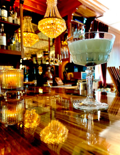 A cocktail glass sits on a polished bar, reflecting warm light from chandeliers and candles in a cozy setting.