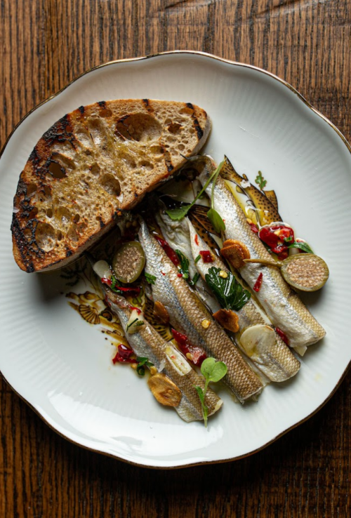 A plate with grilled fish, garnished with herbs and sauces, alongside a slice of toasted bread.
