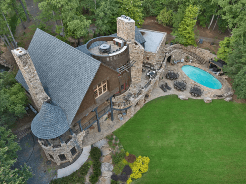 Aerial view of a stone house with a circular tower, surrounded by greenery and a pool in the backyard.