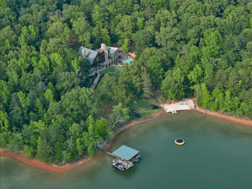 Aerial view of a lakeside home surrounded by lush greenery, with a dock and a floating water toy on the water.