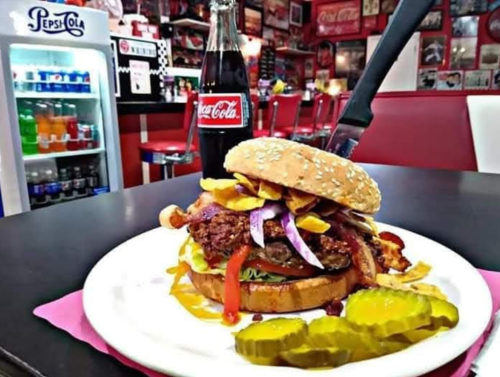 A large burger with toppings, served on a plate with pickles, alongside a bottle of Coca-Cola in a retro diner setting.