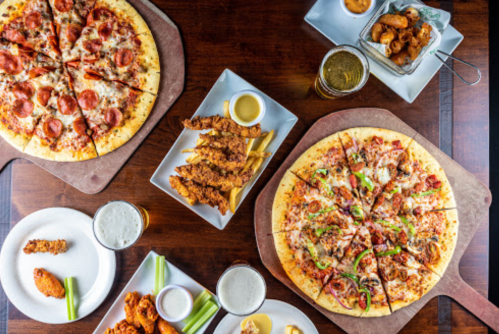 A table spread with two pizzas, chicken tenders, wings, celery, dipping sauces, and drinks.