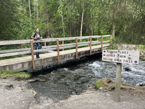 A person stands on a wooden bridge over a stream, with a signpost pointing to nearby attractions in a forested area.