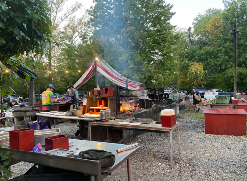 Outdoor cooking area with smoke, grills, and people preparing food under a colorful tent surrounded by trees.