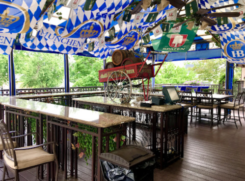 A decorated outdoor bar with a stone countertop, beer banners, and a vintage cart, surrounded by greenery.