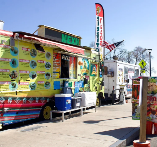 Colorful food truck with a menu of Mexican dishes, parked beside another truck, on a sunny day.