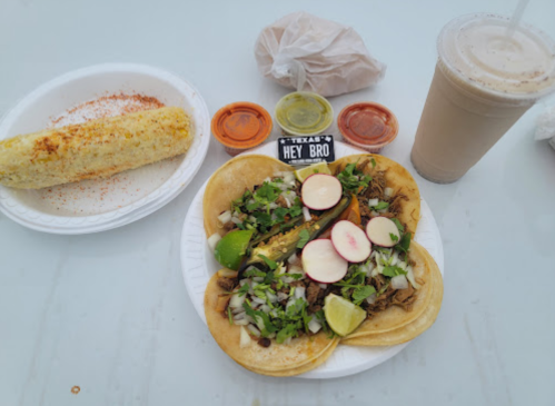 A plate of tacos with meat, onions, and lime, accompanied by corn on the cob and drinks, with sauces on the side.