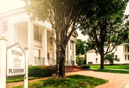 A charming white building with columns, labeled "Weston Playhouse," surrounded by greenery and trees.