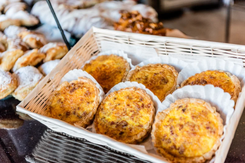 A basket of six golden-brown quiches on white paper liners, with assorted pastries in the background.