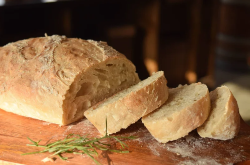 A freshly baked loaf of bread on a wooden cutting board, with a few slices cut and a sprig of herbs nearby.