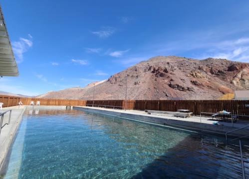 A clear swimming pool surrounded by a wooden fence, with a rocky mountain backdrop under a blue sky.