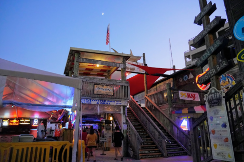 A lively outdoor dining area with colorful signs, stairs, and a tent, set against a twilight sky.