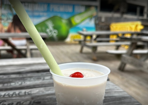 A refreshing drink with a cherry on top and a green straw, set on a wooden table with a beachy background.