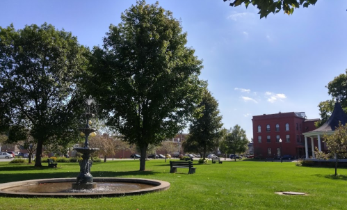 A sunny park scene featuring a fountain, green grass, trees, and a red building in the background.