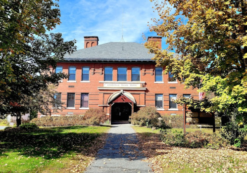 Historic red brick school building surrounded by trees and autumn foliage under a clear blue sky.