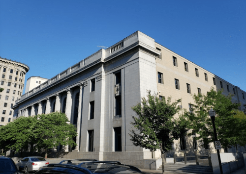 A large, historic building with columns and a modern facade, surrounded by trees and clear blue sky.