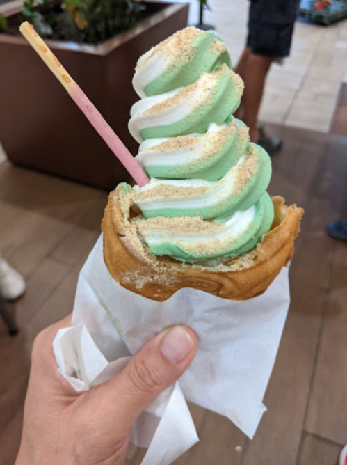 A hand holding a swirl of green and white soft serve ice cream in a waffle cone, topped with a sprinkle of cinnamon.