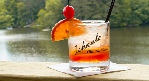 A refreshing Old Fashioned cocktail garnished with a cherry and orange slice, set against a scenic lake backdrop.