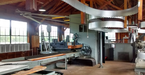 Interior of a woodworking shop featuring large machinery, saw blades, and wooden beams, with natural light from windows.
