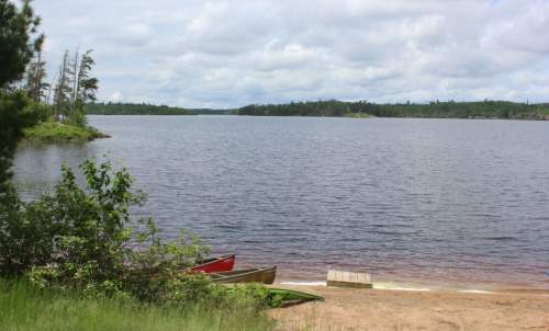 A serene lakeside view with canoes on the shore, surrounded by trees and a cloudy sky.