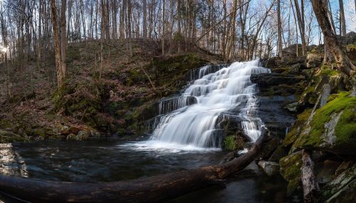 A serene waterfall cascades over rocks, surrounded by trees and moss, with a tranquil pool at its base.