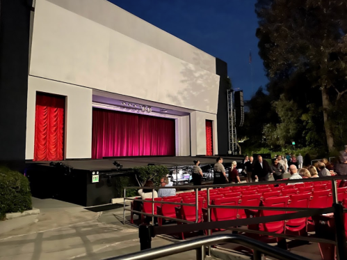An outdoor theater with red curtains, empty red seats, and a few people gathering before a performance at dusk.
