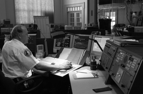 A police officer monitors multiple screens in a control room, focused on data and communications in a black and white setting.