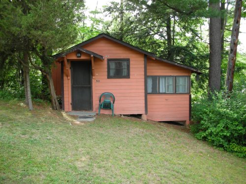 A small, orange cabin surrounded by trees, with a green chair on the porch and a grassy area in front.