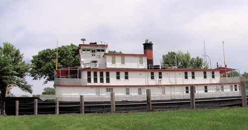 A large, historic riverboat with a red and white exterior, docked near a grassy area and surrounded by trees.