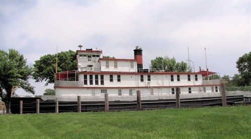 A large, white and red riverboat docked on land, surrounded by green grass and trees under a cloudy sky.
