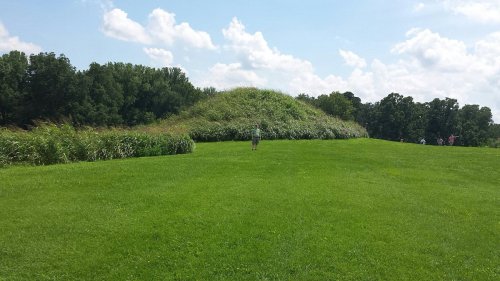 A person stands on a grassy field in front of a large, grassy mound under a blue sky with scattered clouds.