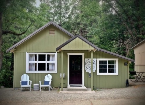 A charming green cottage with a welcoming porch and two white chairs, surrounded by trees.