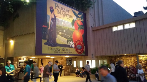 A mural of a woman in a gown with oranges, titled "Pageant of the Masters," at Laguna Beach, California, with people walking by.