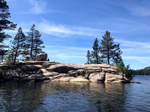 A rocky shoreline with trees and a clear blue sky reflected in calm water.