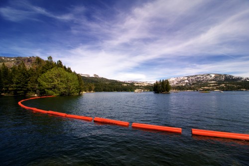 A serene lake scene with a bright orange barrier floating on the water, surrounded by trees and mountains under a blue sky.