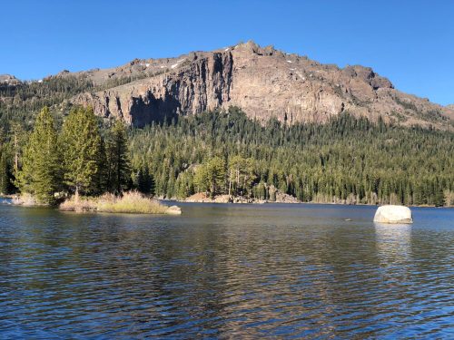 A serene lake surrounded by trees, with a rocky mountain in the background under a clear blue sky.